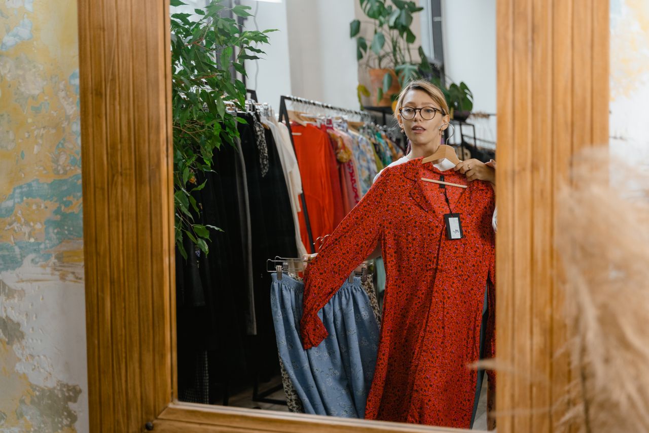 A woman stands in a boutique, viewing a red dress in a mirror, showcasing fashion choices.